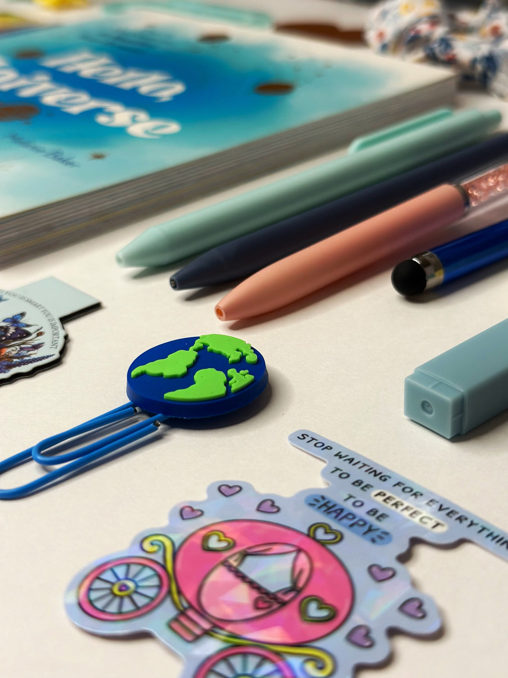 A styled flat-lay of various stationery items on a light-colored desk, including a blue paperclip with a globe design, a pink and blue sticker with a carriage and the text 'STOP WAITING FOR EVERYTHING TO BE PERFECT TO BE SHAPED', pastel pens, and a notebook.
