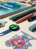 A styled flat-lay of various stationery items on a light-colored desk, including a blue paperclip with a globe design, a pink and blue sticker with a carriage and the text 'STOP WAITING FOR EVERYTHING TO BE PERFECT TO BE SHAPED', pastel pens, and a notebook.